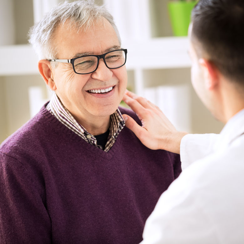 An older man in glasses and a purple sweater smiles and looks at a younger male doctor who puts his hand on the older man's shoulder.