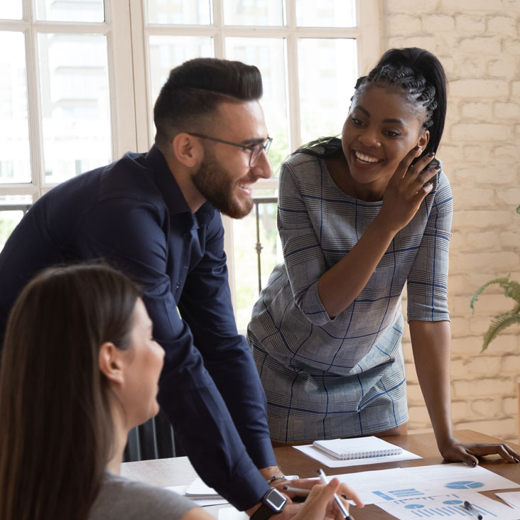 A diverse group of coworkers meet, talk and laugh at a conference table looking at print-outs of charts and graphs.