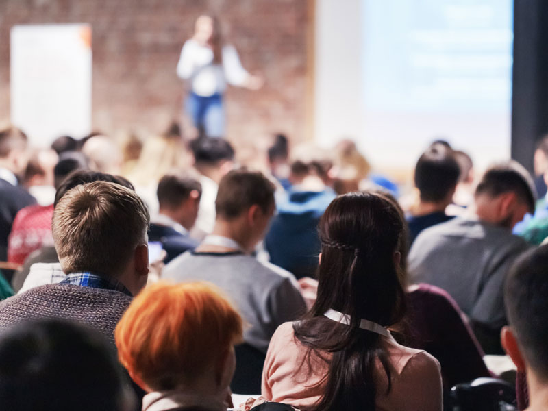 A large, diverse group of young people with their backs to the camera attending a speaking event hosted by a woman on stage who is out of focus.