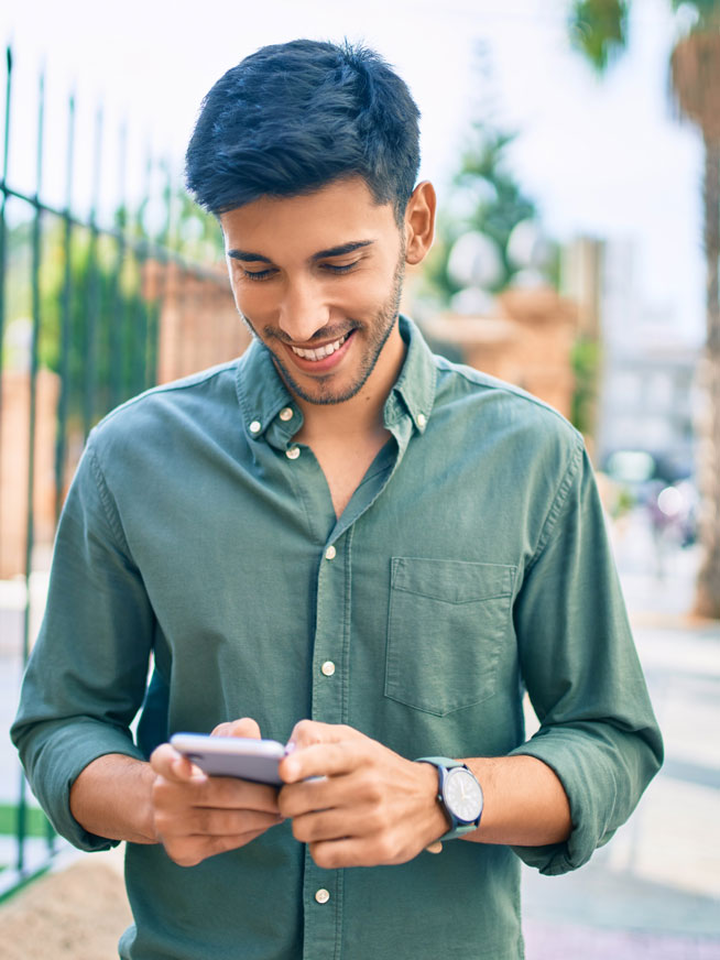 A young man in green walks down a busy suburban street while smiling at the black smartphone in his hands.