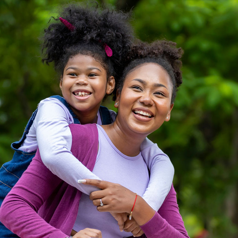 A young girl in overalls climbs on her mom's back, who is wearing a purple jacket, while both smile and look to their right on a blanket in a green pasture.