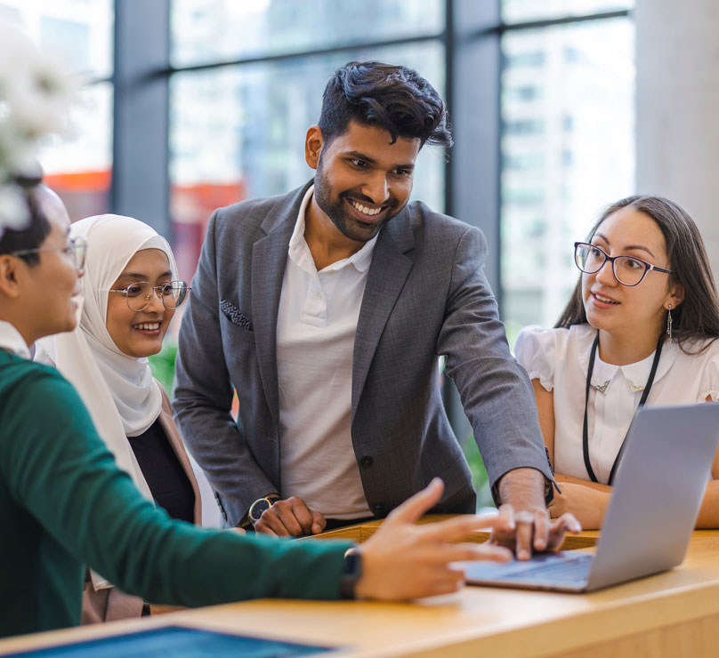 Four people sit at a table in a modern office and share a single laptop, smiling and conversing with one another.