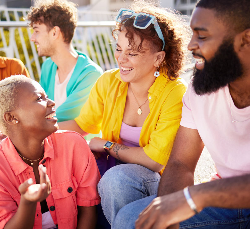 A diverse group of friends in bright colors sit around each other on a sunny patio, smiling and laughing.