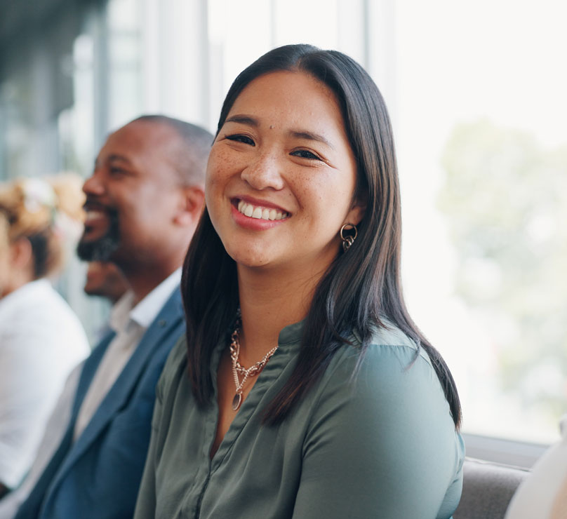 A small group of people sit in a row in a modern office. The person in focus is a young woman in green with dark hair smiling directly at the camera.
