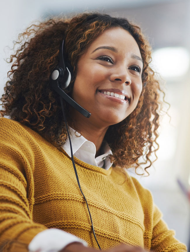 A young female receptionist with a yellow collared shirt and a black headset smiles and receives a phone call while using her computer.