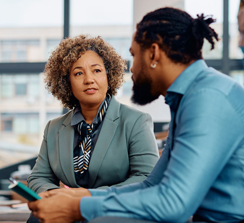 A group of people attend a large, modern lecture hall. In the foreground, a man in blue holding a notebook talks to woman in a grey suit.