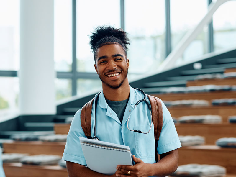 A young male medical student wearing scrubs and a stethoscope while carrying a backpack in a modern, wooden lecture hall.