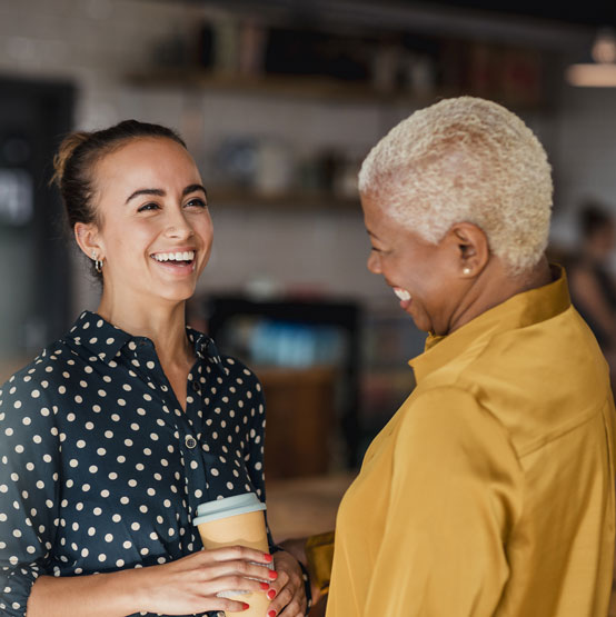 Two young professional women, one win blue with white dots and the other in yellow, talk at a coffee shop and smile at one another.