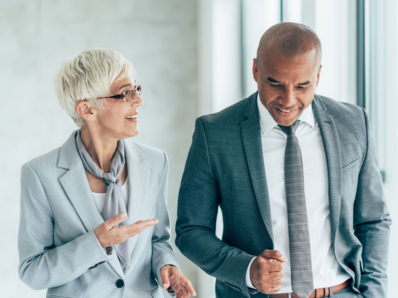 A professional man and women, both in grey business suits, smile and converse while walking on stairs.