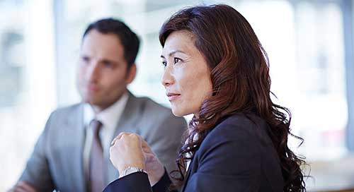 A professional woman in a suit sits and listens intently in a professional office setting with a man out of focus in a grey suit sits in the background