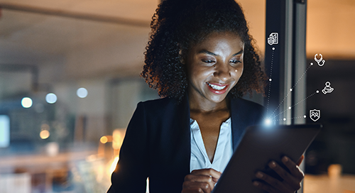 A woman in professional attire in the evening smiles and works on a tablet device, which illuminates her face
