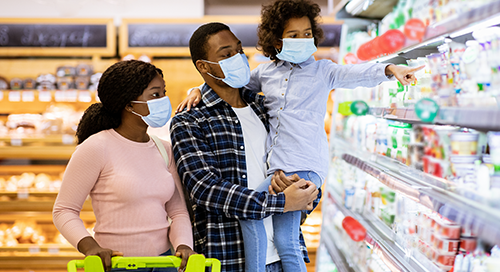 A family of three wearing medical masks shop for groceries as the father holds the child and the mother pushes the cart.