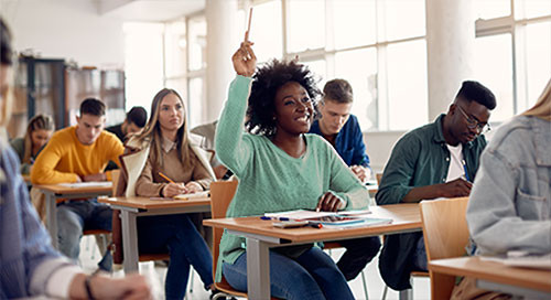 A classroom is filled with diverse students as a female student in green smiles and raises her hand while holding a pencil