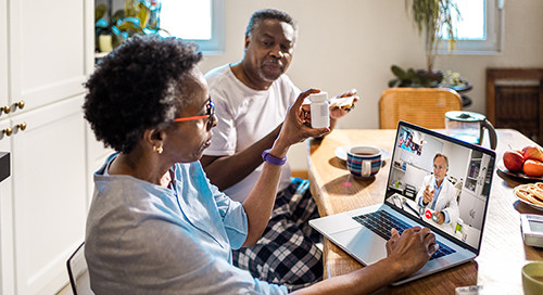 An older couple sitting at their kitchen table hold up a pill bottle while on a digital conference with a doctor in a white coat