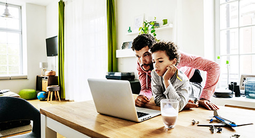 A father and son work in a home office setting on a shared laptop computer