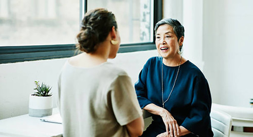 Two woman, one in blue and one in grey, sit in an office setting and converse with one another