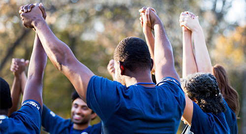 A diverse group of people in matching blue shirts hold hands and cheer outside