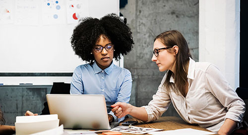 Two professional women, one in blue and one in white, sit around a conference table speaking to one another and gesturing toward a laptop computer