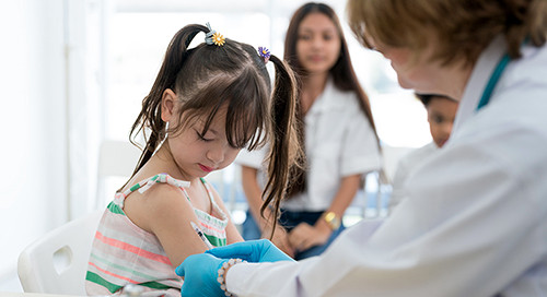 A young girl in a striped shirt receives a shot from a medical professional in a white coat and blue gloves as her family watches on in the background