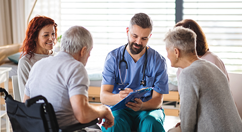 A group of four individuals, including one in a wheelchair, sit in a circle and speak to a medical professional in scrubs as he writes on a clipboard