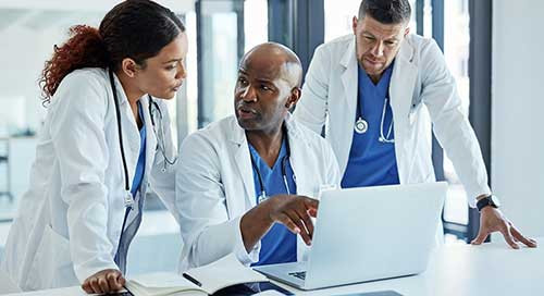 A diverse group of doctors, all in white coats and blue scrubs with stethoscopes, talk to one another and look at a laptop computer