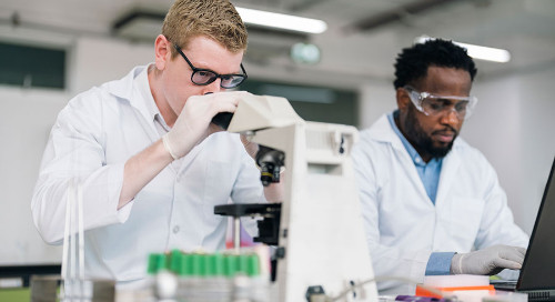Two scientists work at a research table in white coats, gloves, and safety glasses while one uses a laptop and the other uses a microscope