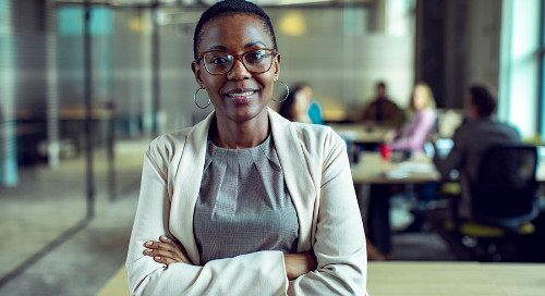An African-American business woman stands in the foreground in beige, smiling with her arms crossed. In the background and out of focus is a modern business office populated with workers.