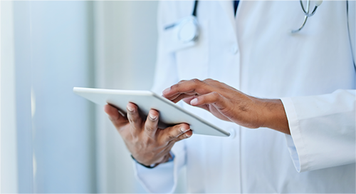 A doctor in a white coat and stethoscope holds and works on a tablet device