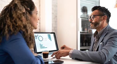 A woman in blue and a man in a grey suit sit at a desk and look at a computer screen with graphic data