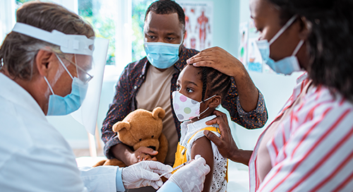 A young girl with her parents, all wearing medical masks, receives an injection from a doctor in a white coat, blue mask, and clear facial screen in a medical office setting