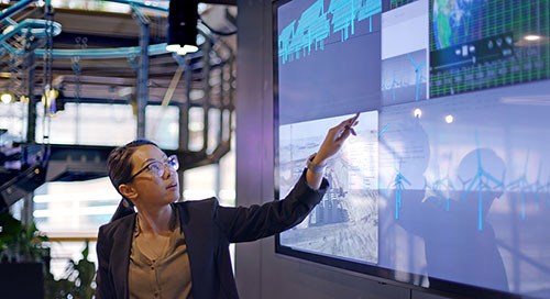 A woman in a brown shirt and grey jacket in a conference space gestures toward a wall of interactive TV screens