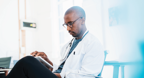 A doctor with glasses, a white lab coat, and stethoscope sits in a professional setting while working on a tablet device