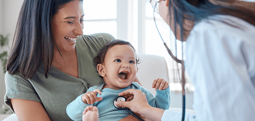 A smiling infant in blue is held by its mother in green who is also smiling as a medical professional with a stethoscope and a white coat listens to the child's heartbeat