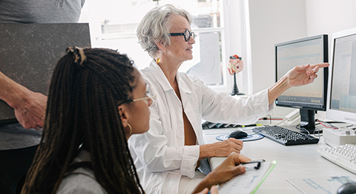 Two women, one older and one younger, converse and gesture toward computer screens