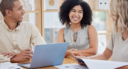 Three people sit around a table strewn with papers as they smile at one another and work at a laptop computer