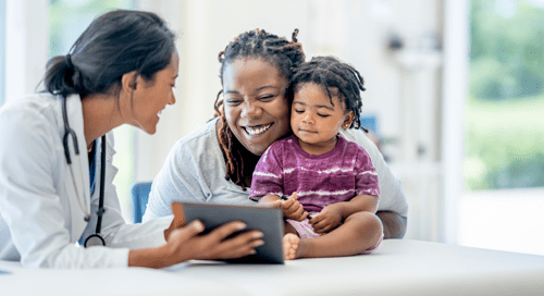A smiling doctor with a white coat, blue scrubs, and a stethoscope holds out a tablet for a woman and child to look at - the woman in grey and the child in purple