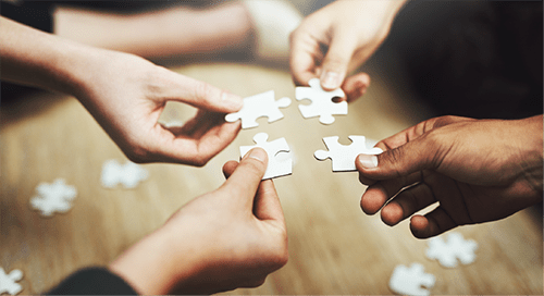Four diverse hands hold out four white puzzle pieces as they sit in a circle