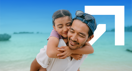 A man in a white shirt and denim cap smiles while his daughter rides on his shoulders at the beach, with the Gainwell arrow logo in the background