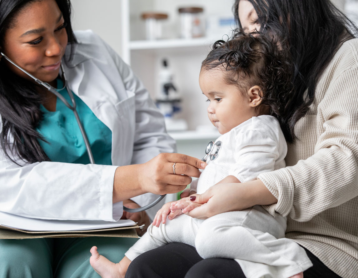 A toddler sits in her mother's lap while a smiling doctor holds a stethoscope up to her heart.