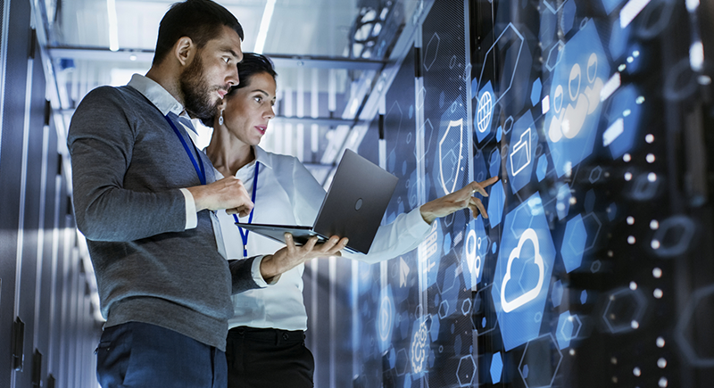 A woman and and a man in professional attire are in a hypermodern server room, gesturing at a large screen with tiles and icons on it while the man holds up a laptop computer.