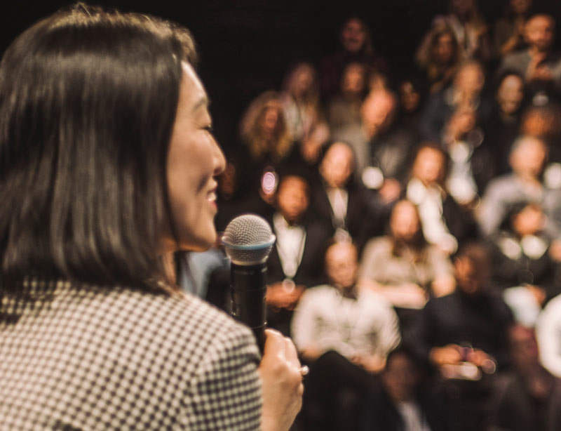 A woman smiles and holds a microphone on stage while an audience is out of focus in the background at a conference event