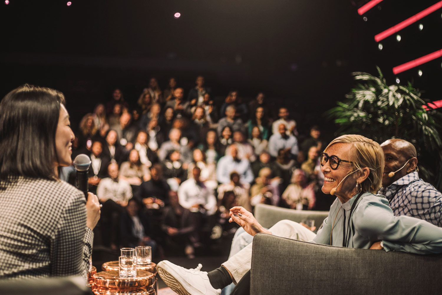 Two women and a man sit on a stage with microphones, smiling and speaking with one another while a conference audience looks on listening