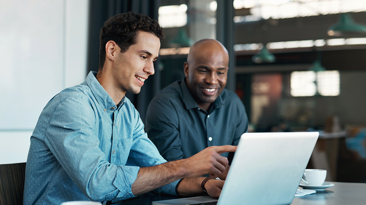 Two men collaborate side-by-side in front of a laptop at a table in a modern office exploring ways to modernize Medicaid.
