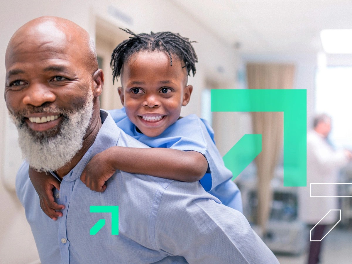 An African-American man with a white beard and blue shirt carries a young child in a medical gown smiling on his shoulders while a doctor and nurse are out of focus in the background