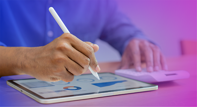 An individual in a blue shirt holds a digital stylus while using a tablet with charts and graphs while also using a digital calculator
