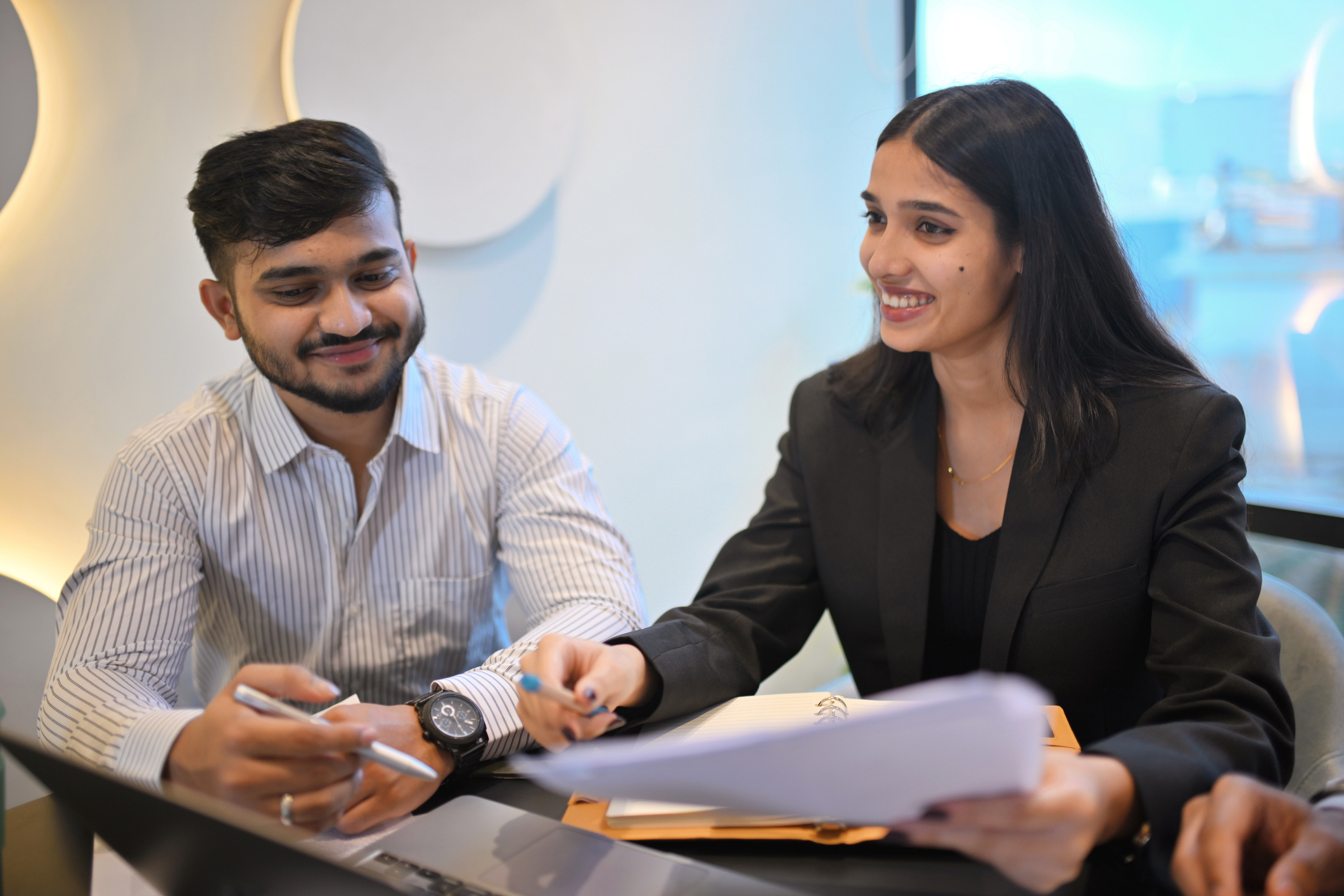 A male and female colleague sit at a table in a meeting. The woman is presenting with a paper in one hand while gesturing with the other.